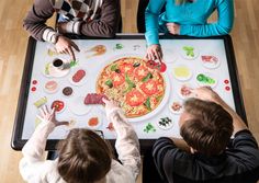 three people sitting at a table with a large pizza on the center piece and toppings