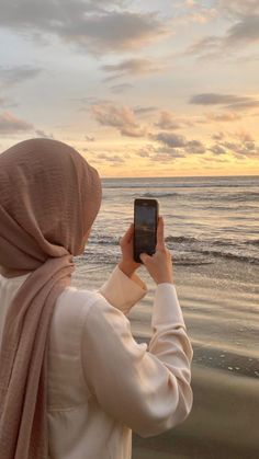 a woman wearing a hijab is looking at the ocean with her cell phone