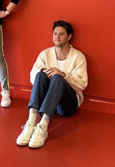 a man sitting against a red wall with his feet up on the ground and wearing white sneakers