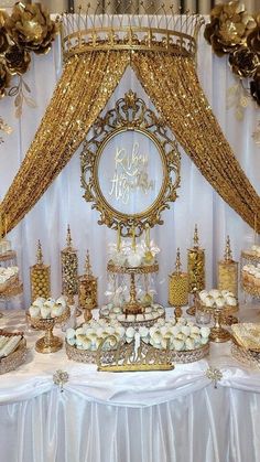 a table topped with lots of cakes and desserts next to a gold drape