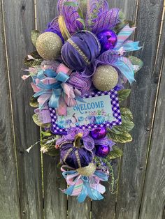 a wreath with purple and blue decorations hanging on a wooden fence next to a sign that says welcome