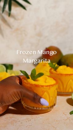 a person holding a small glass filled with mango margaritas on top of a counter