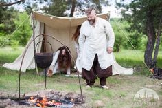 a man standing in front of a fire next to a tent with a cooking pot on it