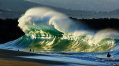 two people are standing on the beach near large waves in the ocean and one person is swimming