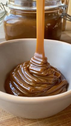 a spoon full of caramel sauce in a white bowl on a wooden table next to jars