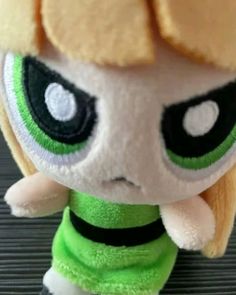 a close up of a stuffed animal toy with big eyes and blonde hair, wearing a green shirt