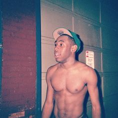 a shirtless man standing in front of a red brick wall wearing a green hat