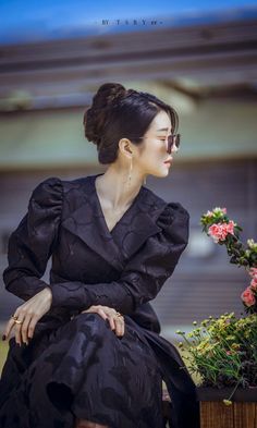 a woman in black dress sitting next to flowers and potted plant with her eyes closed