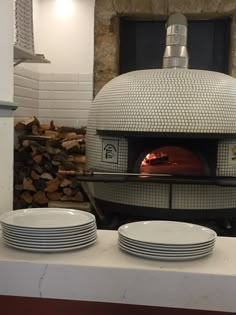 a pizza oven with plates stacked in front of it and stacks of wood behind it