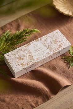 a white box sitting on top of a table next to green leaves and pine branches