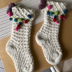 two white knitted stockings with colorful pom - poms on them sitting on a table