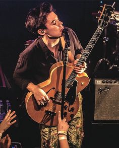 a man holding a guitar while standing next to a microphone and other people in front of him