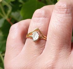 a woman's hand holding a ring with a white stone in the middle and green leaves behind it