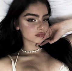 a woman laying on top of a bed wearing a white shirt and pearl necklace with her hand under her chin