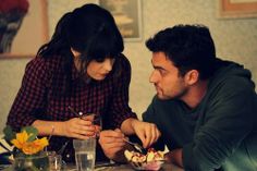 a man and woman sitting at a table with food in front of them, looking at each other