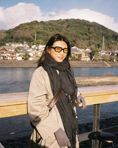 a woman standing on a bridge with her hands in her pockets, wearing sunglasses and a scarf