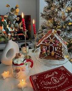 a table topped with a gingerbread house next to a christmas tree and lit candles