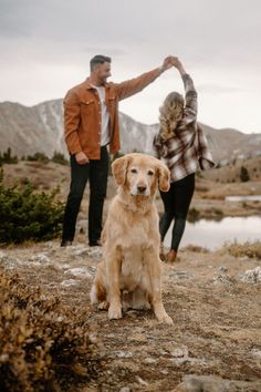 a man and woman standing on top of a mountain holding hands with a golden retriever