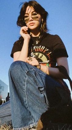 a woman sitting in the grass with her hand on her chin, wearing sunglasses and holding a black bag