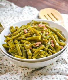 a white bowl filled with green beans on top of a floral table cloth next to a wooden spoon