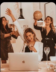 three women at a table with wine glasses in front of a laptop and one holding a wooden ball above her head