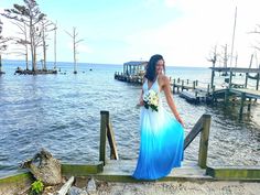 a woman in a blue and white dress standing on a dock by the water with her bouquet