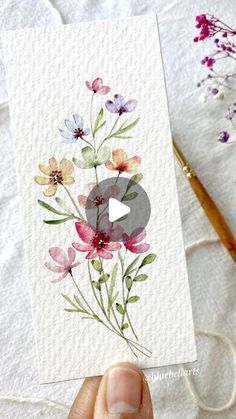 someone holding up a card with watercolor flowers on it and yarn in the background