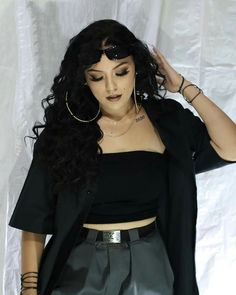 a woman with long black hair wearing a crop top and grey pants, standing in front of a white backdrop