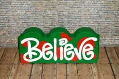 a wooden sign with the word believe painted on it in red and green, against a brick wall