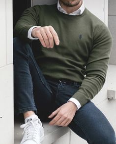 a man in a green sweater and jeans leaning against a wall