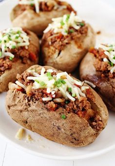 several baked potatoes on a white plate topped with cheese and chili sauce, sprinkled with green onions