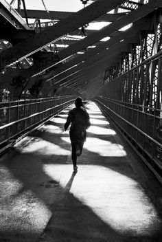 black and white photograph of person running on bridge with sun shining through the metal beams