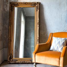 an orange chair sitting in front of a large gold framed mirror next to a window