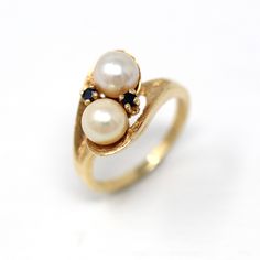 Classic vintage circa 1960s retro era 14k yellow gold cultured pearl ring! This stylish Toi et Moi style ring features two round cultured pearls, flanked by sapphire accents. The setting has a timeless bypass design with a pretty brushed finish. A timeless piece of fine retro era jewelry, featuring June & September's birthstone! *Sale - price reduced from $395 USD to $385 USD.  ERA - Circa 1960s - Retro METAL / MATERIAL - 14k yellow gold, 2 genuine 6 mm cultured pearls, two sapphires MARKINGS / HISTORY - Inside of band is marked 14k with a maker's mark in a triangle  CONDITION - Good vintage condition. Yellow gold metal has been professionally polished & cleaned. Age appropriate patina & wear remains. Cultured pearls are secure. Amazing vintage cultured pearl ring!   SIZE / MEASUREMENTS - Vintage Multi-stone Pearl Ring For Anniversary, Vintage Multi-stone Pearl Ring For Formal Occasions, Vintage Multi-stone Pearl Ring For Formal Events, Vintage Pearl Ring With Gemstone, Vintage Yellow Gold Pearl Ring With Gemstone, Vintage White Pearl Ring, Vintage Pearl Ring With Gemstone For Formal Occasions, Gold Multi-stone Pearl Ring For Anniversary, Heirloom Multi-stone Pearl Ring For Anniversary