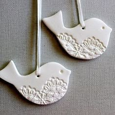 two white ceramic birds hanging from strings on a gray tablecloth, one is decorated with lace and the other has an ornament in the shape of a bird