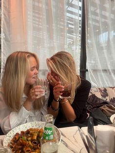 two women sitting at a table with food and drinks in front of them, drinking wine