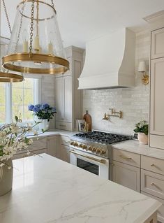 a white kitchen with marble counter tops and gold accents on the hood, windows, and light fixtures