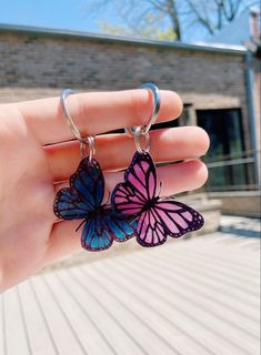 a person holding two purple and blue butterflies on their earring hooks in front of a brick building