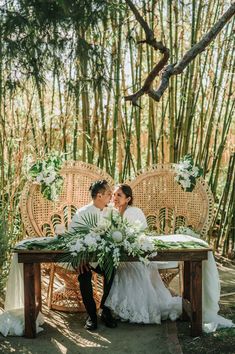 a bride and groom sitting at a table with flowers in front of some bamboo trees