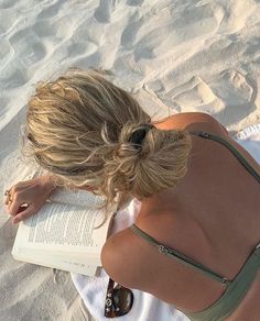 a woman sitting on the beach reading a book with her hair in a pony tail