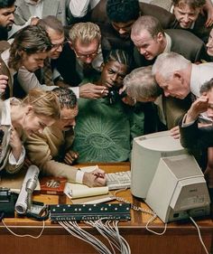 a group of people standing around a computer on top of a wooden desk next to each other