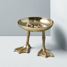 a gold bird shaped bowl with legs and feet on the side, sitting in front of a white wall