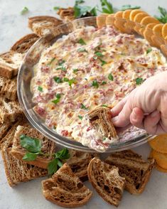 a hand dipping some crackers into a dip in a glass bowl with bread on the side