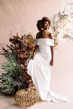 a woman in a white dress standing next to some plants and flowers on a pink background