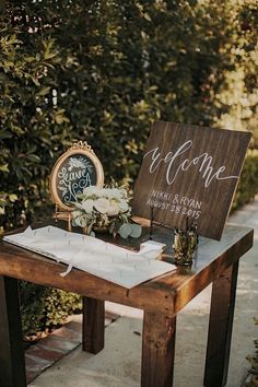 a wooden table topped with a sign next to a clock and vase filled with flowers