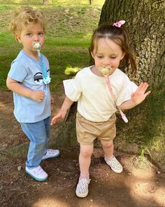 two young children standing next to a tree eating something off of a spoon in their mouth