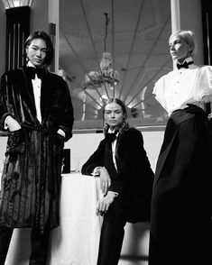 three women standing next to each other in front of a window wearing long dresses and tuxedos