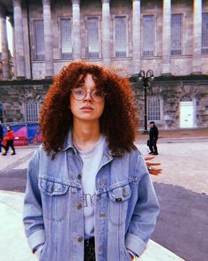 a woman with red curly hair and glasses standing in front of a large stone building