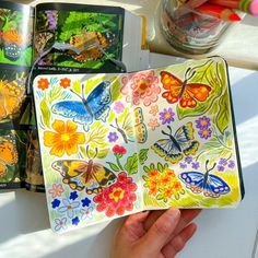 a hand holding an open book with butterflies painted on it and other books in the background