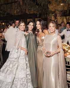 three women in dresses standing next to each other at a dinner table with people around them